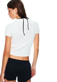Biely, krátky rukáv, pletený polo top s textúrovaným vzorom a čiernym detailom na viazanie vzadu, kombinovaný s čiernymi šortkami.