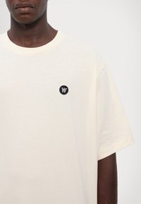 Kremfarget bomull t-skjorte med rund hals og korte ermer, med et lite svart sirkulært merke med en hvit "W"-logo.