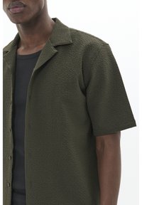 Camisa de manga corta color verde oliva con un patrón texturizado similar a un gofre. Cuenta con cuello tipo rever y cierre frontal con botones.
