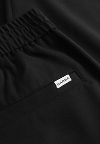 Zwarte broek met een elastische tailleband en een wit label met de tekst "GABBA." De stof lijkt glad zonder zichtbare patronen of accenten.