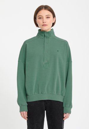 Ung person med kort brunt hår iført en grøn højhalset sweatshirt og sorte bukser mod en ensfarvet hvid baggrund.