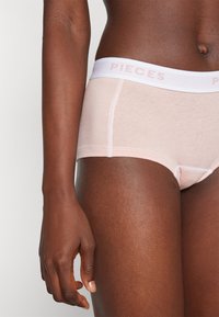 Lys rosa bomullsundertøy med hvit elastisk midjebånd med teksten "PIECES". Stoffet er glatt med en lett glans.