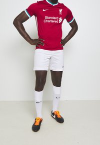 Camiseta roja del Liverpool FC con pantalones cortos blancos. La camiseta presenta acentos azules y el logo del patrocinador; el jugador lleva botas negras con cordones naranjas y calcetines blancos.