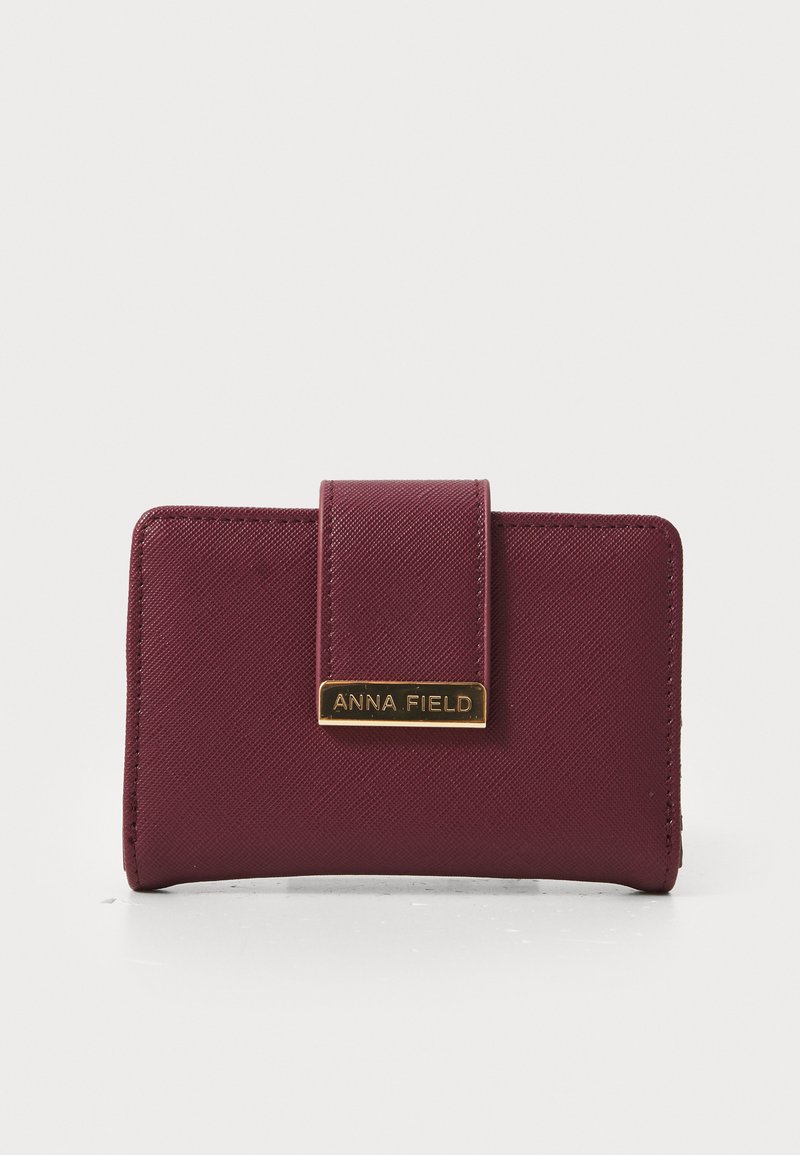 Portofel burgundy cu o suprafață texturată, având o plăcuță logo rectangulară aurie și închidere magnetică. Formă compactă rectangulară.