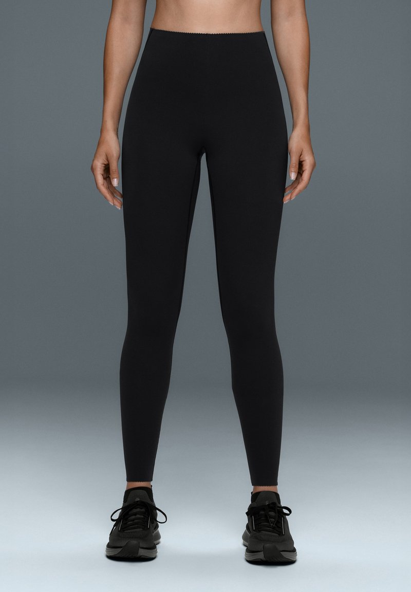 Modèle debout en legging noir taille haute et baskets noires sur fond gris de studio.