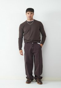 Bruine lange mouwen trui, donkerpaarse wijde broek en bruine veterschoenen met witte stiksels, tegen een witte achtergrond.