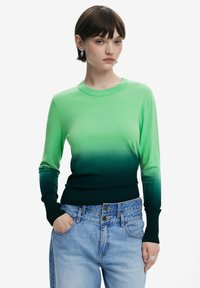 Grüner, gradientfarbener Strickpullover mit Rundhalsausschnitt, langen Ärmeln und gerippten Bündchen, kombiniert mit hellblauen, hochgeschnittenen Jeans.