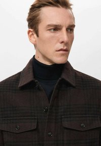 Chaqueta marrón a cuadros hecha de tela suave, con cuello con solapa, cierre de botones y dos bolsillos en el pecho. Se lleva sobre un suéter de cuello alto negro.