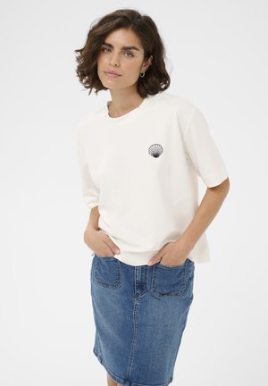 Frau mit kurzen braunen Haaren trägt ein weißes, lockeres T-Shirt mit einem schwarzen Muschelsymbol und einem knielangen blauen Jeansrock, die Hände in den Taschen.