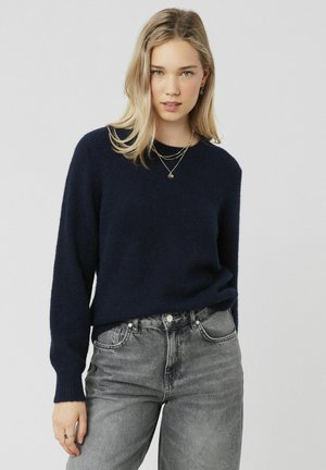 Pull en maille bleu marine avec manches longues, col rond et poignets côtelés, associé à un jean en denim gris taille haute.