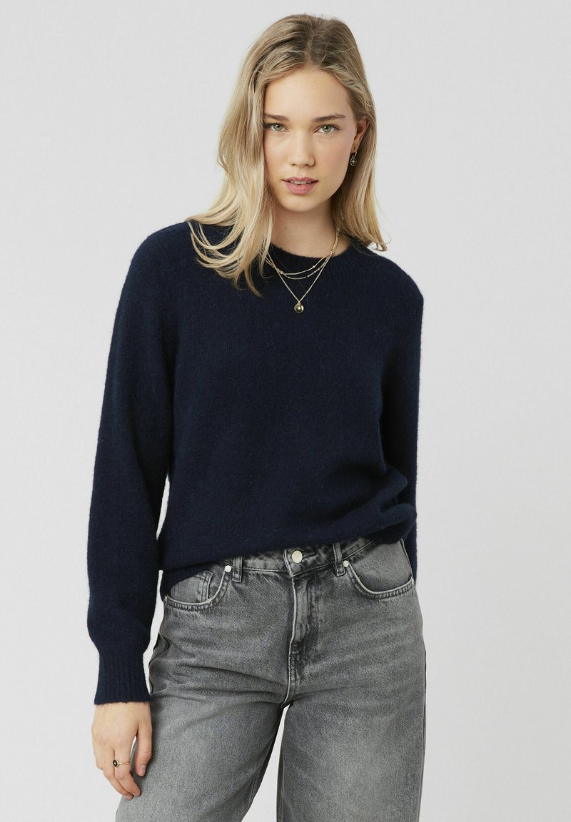 Pull en maille bleu marine avec manches longues, col rond et poignets côtelés, associé à un jean en denim gris taille haute.