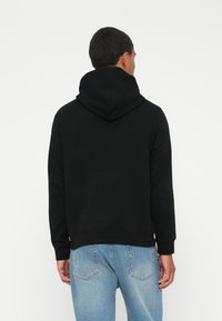 Hombre de espaldas con sudadera negra y jeans azul claro frente a un fondo blanco liso.