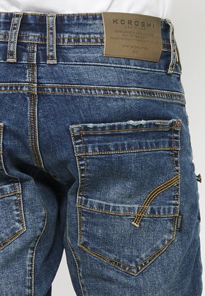 Blaue Denim-Jeans mit einem leicht verwaschenen Finish, ausgestattet mit gelben Nähten, mehreren Taschen und einem markierten, hellbraunen Lederpatch am Bund.