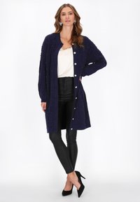 Cardigan lavorato a maglia blu navy con motivo testurizzato e dettagli di perle, caratterizzato da una chiusura con bottoni frontali. Abbinato a leggings in ecopelle neri e tacchi.