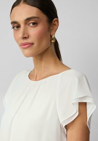 Witte blouse met korte, geplooide mouwen en een ronde halslijn. Model draagt gouden druppeloorbellen. Gladde stof met een lichte glans.