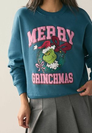 Person trägt einen blauen Pullover mit einer Grinch-Grafik innerhalb eines Weihnachtskranzes und dem Text "Frohe Grinchmas" in pinken Buchstaben.