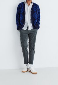 Marineblauwe cardigan met geometrisch patroon, wit overhemd, grijze corduroy broek en witte sneakers met zwarte accenten, tegen een lichte achtergrond.