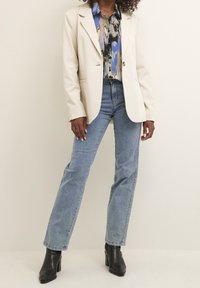 Femme portant un blazer crème, un chemisier bleu à motifs, un jean bleu clair et des bottines à talons noires, debout devant un fond uni.