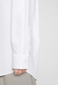 Main partiellement visible portant une manche de chemise blanche à boutons sur un pantalon gris clair sur un fond uni.