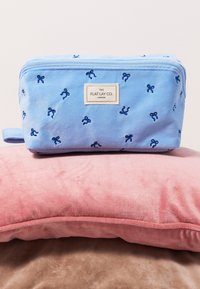 Bolsa de aseo de tela azul claro con bordado de lazo marino y una etiqueta que dice "THE FLAT LAY CO. LONDON", descansando sobre almohadas de terciopelo rosa y beige.