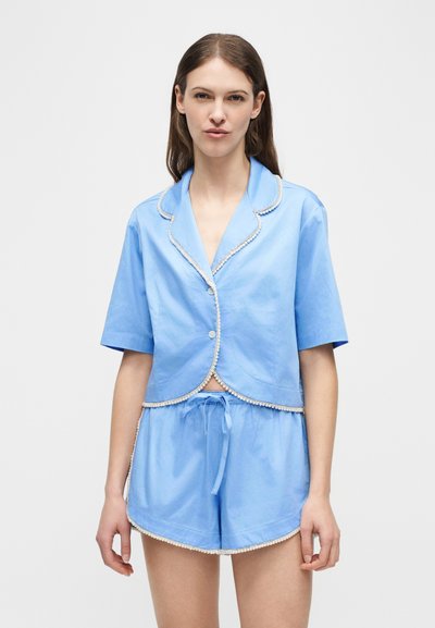 Γυναίκα που φοράει ανοιχτό μπλε μπλουζάκι με κοντά μανίκια και κουμπιά, καθώς και σορτς που ταιριάζουν με άσπρο τελείωμα, στέκεται μπροστά σε απλό ανοιχτό φόντο.