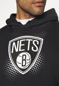 Sweat à capuche noir avec un grand logo blanc des Brooklyn Nets, comprenant un ballon de basket à l'intérieur d'un écusson, agrémenté de motifs graphiques en pointillés.