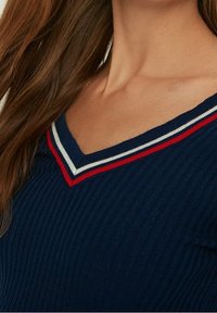 Haut en maille côtelée bleu marine avec un col en V orné de rayures rouges et blanches. Gros plan montrant la texture et les détails du col.
