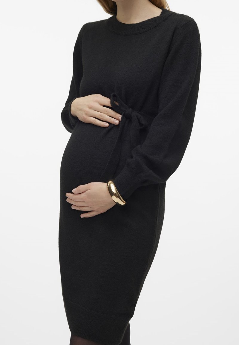 Femme enceinte portant une robe noire à manches longues avec une taille nouée, tenant son ventre des deux mains et portant un bracelet en or.