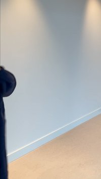 Djelomični prikaz osobe koja nosi tamnu jaknu i stoji pored jednostavnog bijelog zida s bež podom.