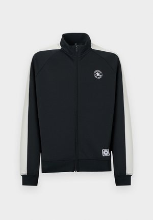 Melna jaka ar rāvējslēdzēju, pelēkām sānu panelēm, ribveida aprocēm un apakšu. Uz krūtīm ir apaļš logotips, priekšpuse ar rāvējslēdzēju.