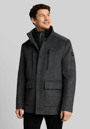 Uomo che indossa un cappotto grigio scuro abbottonato con tasche sul petto con cerniera e tasche con pattina, che guarda leggermente verso l'alto e di lato su uno sfondo semplice.