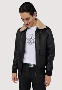 Czarna skórzana kurtka z jasnobeżowym futrzanym kołnierzem, na zamek, z dwiema przednimi kieszeniami, noszona na białej koszulce z graficznym wzorem i ciemnych dżinsach.