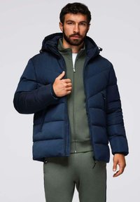 Giacca puffa blu navy con cappuccio, dotata di zip frontale, texture trapuntata e tasche laterali, indossata sopra un zip verde.