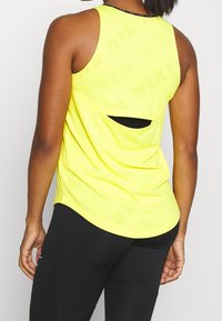 Top atlético sin mangas de color amarillo neón con un corte en la espalda y un sutil patrón de marca. Combinado con leggings negros que cuentan con acentos texturizados.