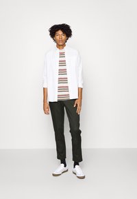 Chemise blanche à manches longues sur un T-shirt rayé beige, marron et vert olive. Pantalon slim olive et baskets blanches avec semelles marron.