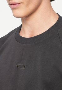 Sweatshirt gris foncé avec un col rond côtelé, des manches raglan et un logo brodé discret sur la poitrine. Texture lisse, design décontracté.