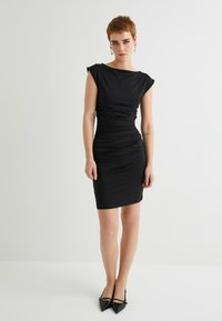 Schwarzes, figurbetontes Kleid mit kurzen Ärmeln, geraffter Textur an der Taille und knielangem Saum. Getragen mit spitz zulaufenden schwarzen Absatzschuhen.