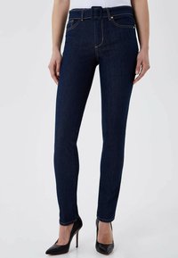 Jean skinny bleu foncé en denim avec une taille haute ceinturée, des coutures visibles et une texture lisse. Associé à des talons hauts noirs.