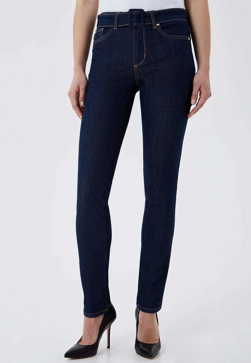 Jean skinny bleu foncé en denim avec une taille haute ceinturée, des coutures visibles et une texture lisse. Associé à des talons hauts noirs.