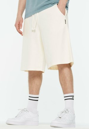 Knielange cremefarbene Shorts aus weichem Stoff mit elastischem Bund und seitlichen Taschen. Kombiniert mit weißen Sneakers und gestreiften Crew-Socken.