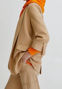 Beige tailored blazer with a relaxed fit, featuring a side pocket and rolled sleeves over an orange hooded sweatshirt. Casual yet refined design.