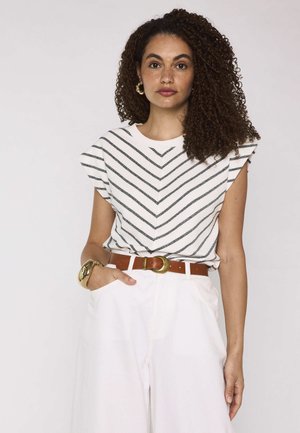 Donna con capelli ricci indossa un top a righe senza maniche, pantaloni bianchi a vita alta con cintura marrone, orecchini a cerchio dorati e braccialetto, posa con la mano in tasca.