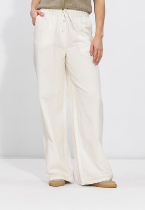 Vrouw draagt losse witte broek met trekkoord, beige trui zonder mouwen en beige instapschoenen, staand tegen een effen achtergrond.