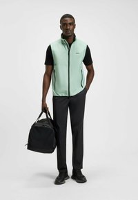 Minzgrüner ärmelloser Weste, schwarzes T-Shirt, schwarze Hose, schwarze Sneakers; hält eine schwarze Reisetasche. Glattes Material mit Reißverschlusstaschen.