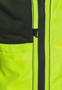 Klart gul jacka med svart nätficka och dragkedjestängning. Har en slät textur och en cirkulär detalj nära fickan.