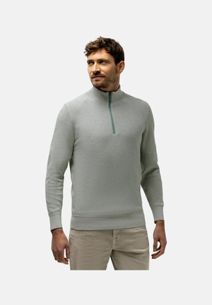 Man wearing a light grey half-zip sweater and beige pants, standing and looking to the side against a plain background.
