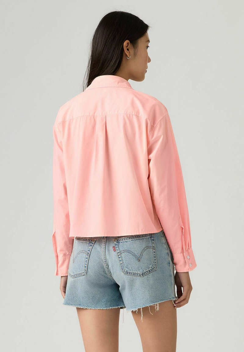 Femme portant un t-shirt à manches longues rose clair et un short en denim bleu clair, vue de dos sur un fond uni.