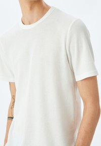 Vit kortärmad t-shirt gjord av strukturerad tyg, med rund hals och ett våffelmönster. Synlig armtatuering nära handleden.