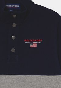 Mörkblå och grå sweatshirt med krage, tre silverfärgade tryckknappar, broderad "POLO SPORT"-logotyp och detalj med USA:s flagga. Slät textur.