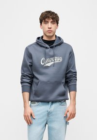 Νεαρός άνδρας που φοράει μπλε φούτερ Converse και ανοιχτόχρωμο τζιν παντελόνι, στέκεται μπροστά σε ένα απλό ανοιχτό φόντο, κοιτάζοντας προς τα εμπρός.